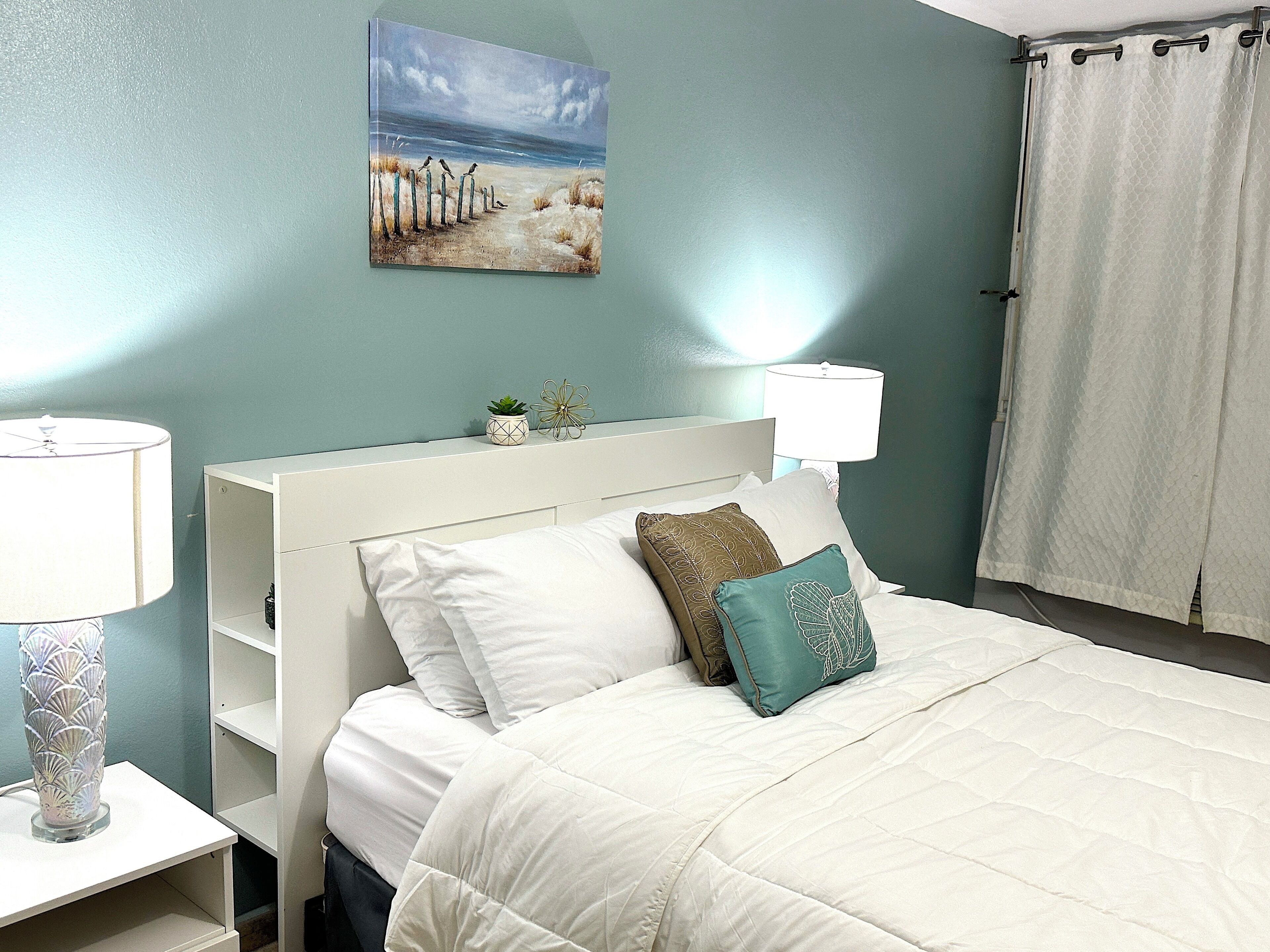 Une chambre à coucher avec un lit, une tête de lit blanche, deux lampes de chevet, une table de chevet, un tableau accroché au mur et une fenêtre avec un rideau.