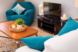 A living room with a teal sofa, a wooden coffee table with a plant, a TV on a dark wooden stand, and a decorative candle.