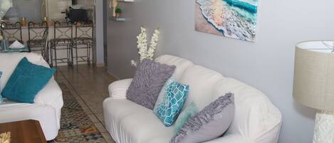 Uma sala de estar com um sofá branco, tapete estampado e uma pintura de uma paisagem praiana.