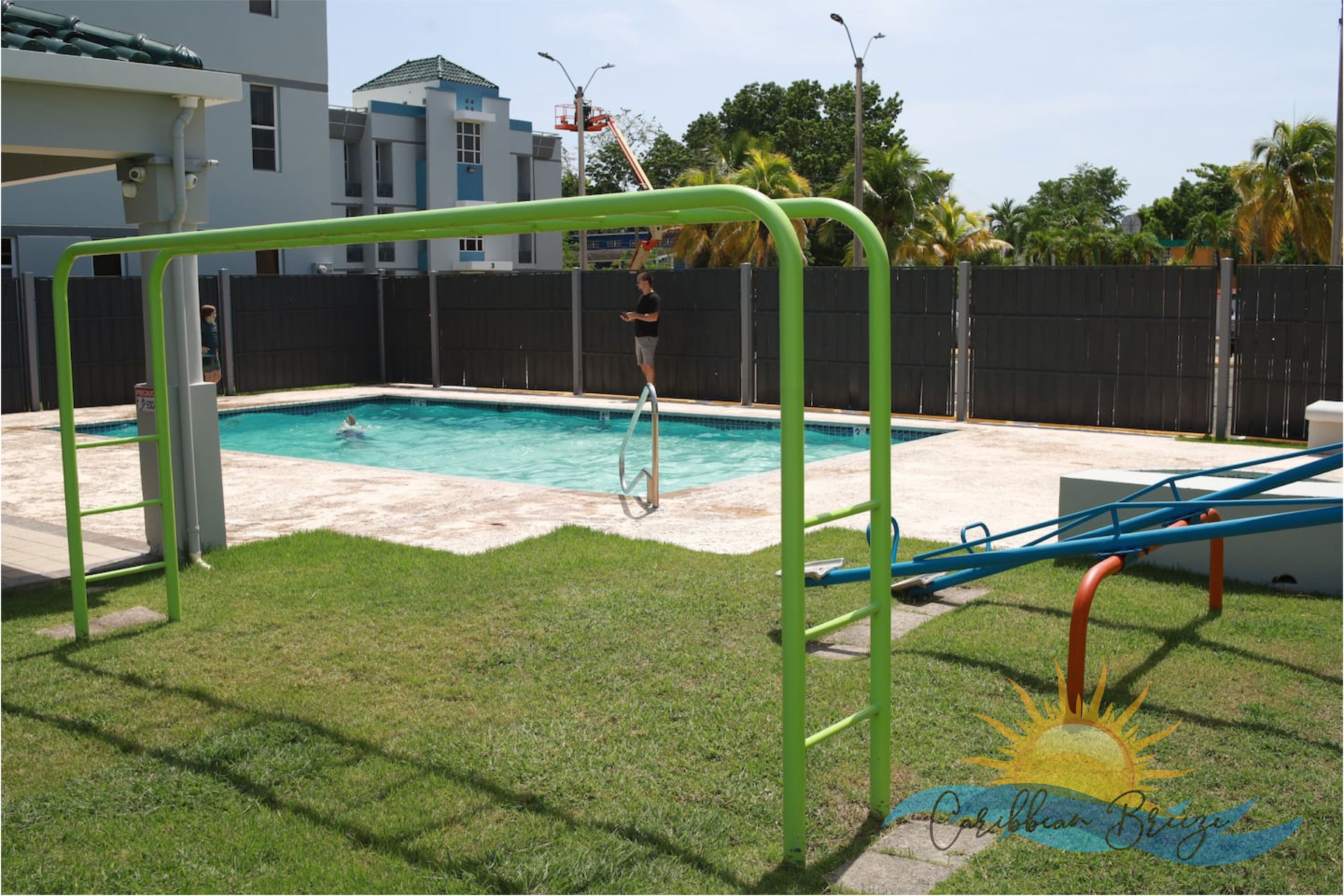 A swimming pool with a green and blue slide, surrounded by a fenced yard and a grassy area.