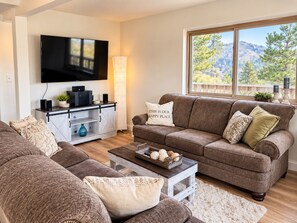 A living room with a brown sofa, a coffee table with decorative items, a TV mounted on the wall, and a window with a view of trees. - Lake View Casa (Fort Collins)