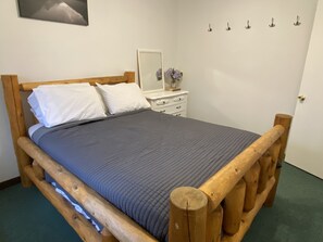 Un lit en bois avec une couverture grise, une table de chevet blanche avec un miroir et un vase de fleurs, et un mur muni de crochets.