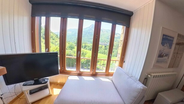 Ett rum med ett stort fönster som erbjuder utsikt över grönskande kullar och ett berg. Det finns en vit soffa och en platt-TV på ett stativ.