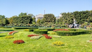 A well-maintained park with manicured lawns, flower beds, and a classical building in the background.