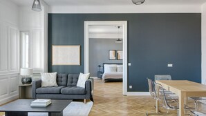 A modern living room with a grey sofa, a wooden coffee table, and a dining area with a wooden table and clear chairs.