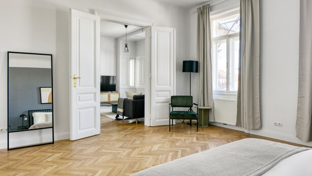 A spacious hotel room with a large bed, a sitting area with a chair and a small table, a large mirror, and a view of the city through the window.