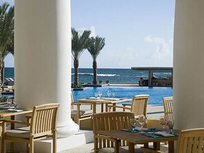 Un espace repas aménagé au bord de la piscine, avec des tables et des chaises en bois, offrant une vue sur la piscine et l’océan.