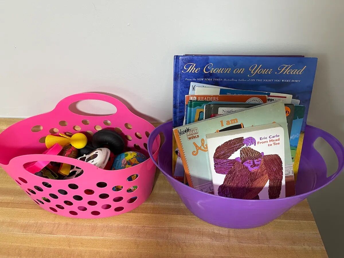 Zwei Körbe auf einer Holztischplatte, einer rosa und einer lila, gefüllt mit verschiedenen Gegenständen wie Büchern und Spielzeug.