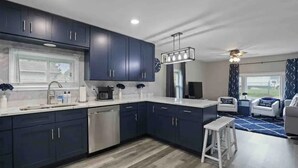 Een moderne keuken met donkerblauwe kastjes, een roestvrijstalen vaatwasser en een wit aanrecht. Er is een bar met een witte barkruk en een plafondventilator.