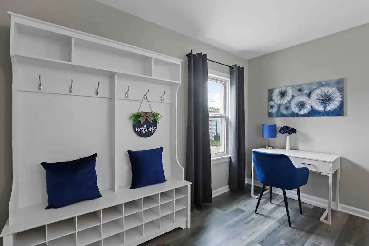 A room with a white bench with blue cushions, a desk with a chair, and a window with curtains.