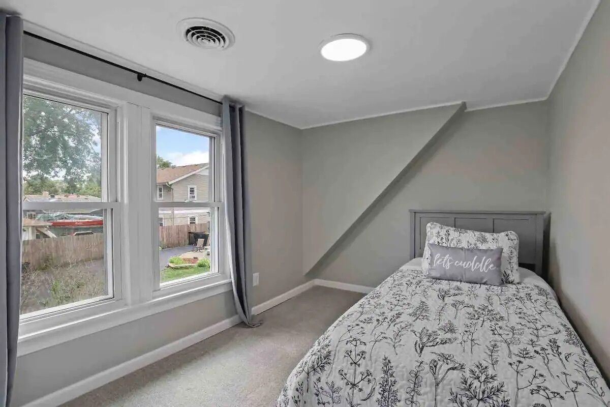 A bedroom with a large window, a bed with floral bedding, and a headboard with a pillow.