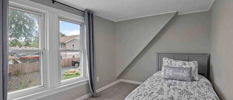 A bedroom with a large window, a bed with floral bedding, and a headboard with a pillow.