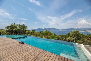 Una piscina infinita de color azul claro con cubierta de madera, rodeada de vegetación exuberante y con vistas a un paisaje costero.