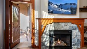 A living room with a stone fireplace, a television mounted on the wall, and a door with wooden blinds. - Village at Palisades Tahoe Condo #6, in Building 5, Ski In/Out, with Mountain View!! (Olympic Valley)