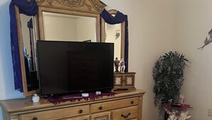 Quarto com uma grande cômoda de madeira, televisão de tela plana, cama rosa, tapete estampado e uma planta decorativa.