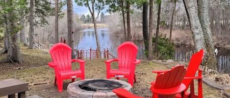 Feuerstelle mit roten Adirondack-Stühlen und einem Steinkamin, umgeben von Bäumen und Blick auf einen See.