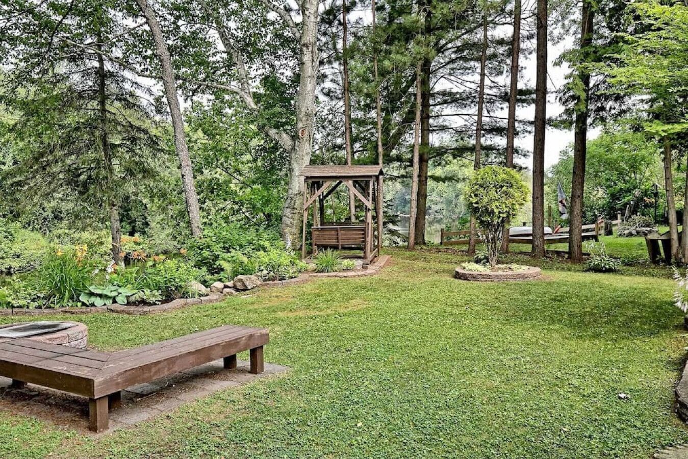 Een houten prieel in een weelderige tuin met een bankje en een kleine boom in het midden.