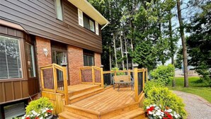A wooden deck with a bench and a chair, flanked by two planters with flowers. - Whispering Pines Riverside Getaway (Kawartha)
