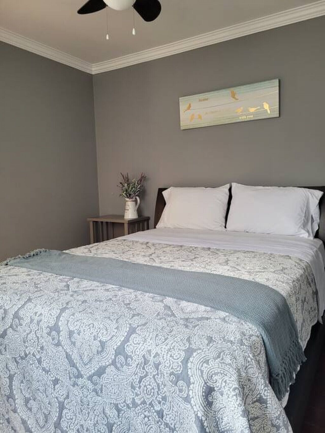 Un dormitorio con una cama grande, una mesita de noche, un ventilador de techo y una obra de arte colgada en la pared.