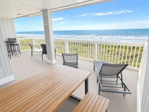 Eine überdachte Terrasse mit einem Holztisch und -stühlen, einem weißen Geländer und Blick auf das Meer.