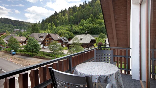A balcony with a table and chairs, overlooking a residential area with houses and a forested hill. - Cozy vacation apartment in spa town in the southern Black Forest with mountain view, garden & Wi-Fi (Todtmoos)