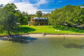 A two-story house with a large deck and solar panels on the roof, surrounded by lush greenery and a sandy beach. - The Punt House - riverfront ramp access (Dunbogan)