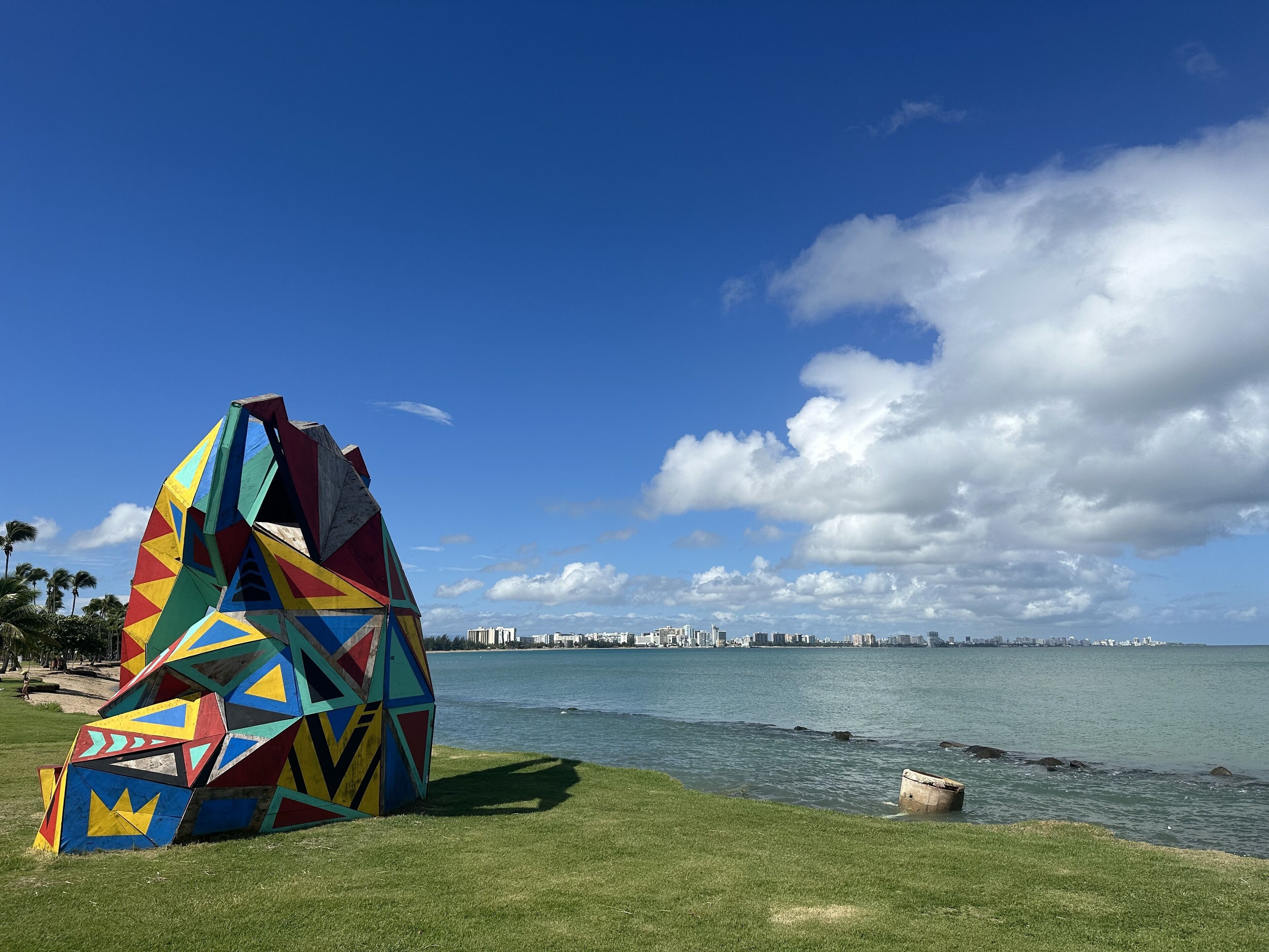 Una escultura geométrica colorida junto al agua, con el horizonte de una ciudad al fondo.