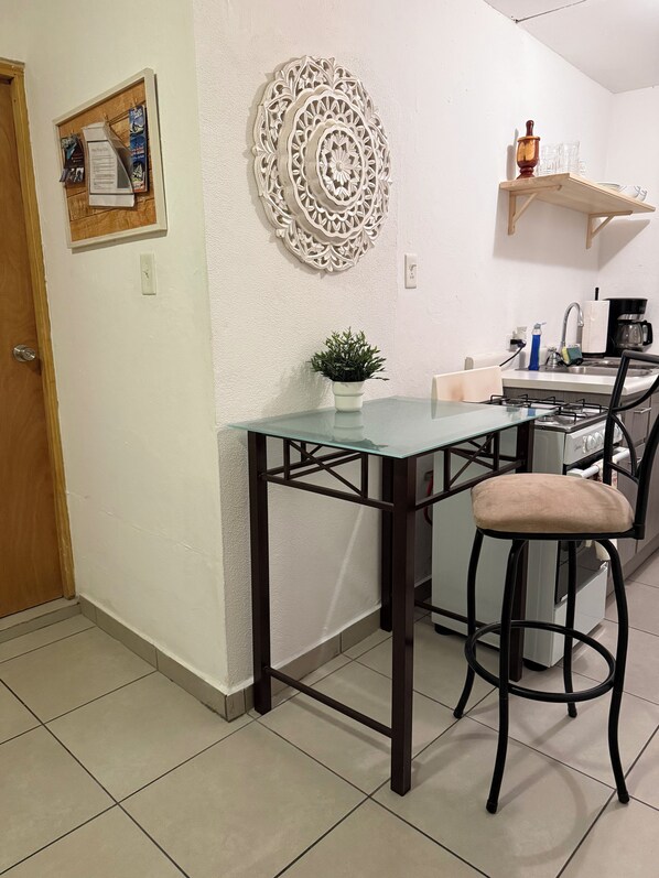 Una cocina con una mesa de vidrio, un taburete de bar, un adorno en la pared y un estante con objetos.