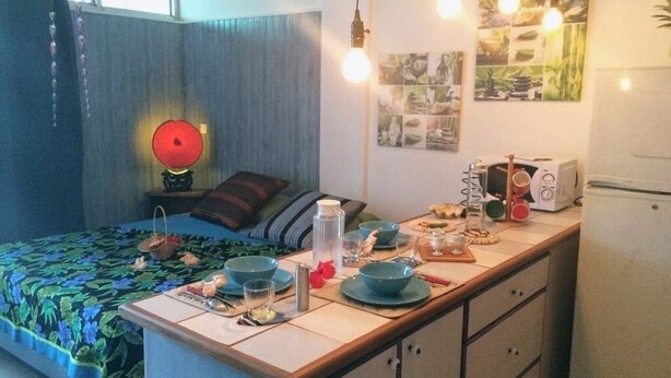 A compact kitchen with a dining area, a bed with a floral bedspread, and a refrigerator. - Ninamu Studio (Papeete)