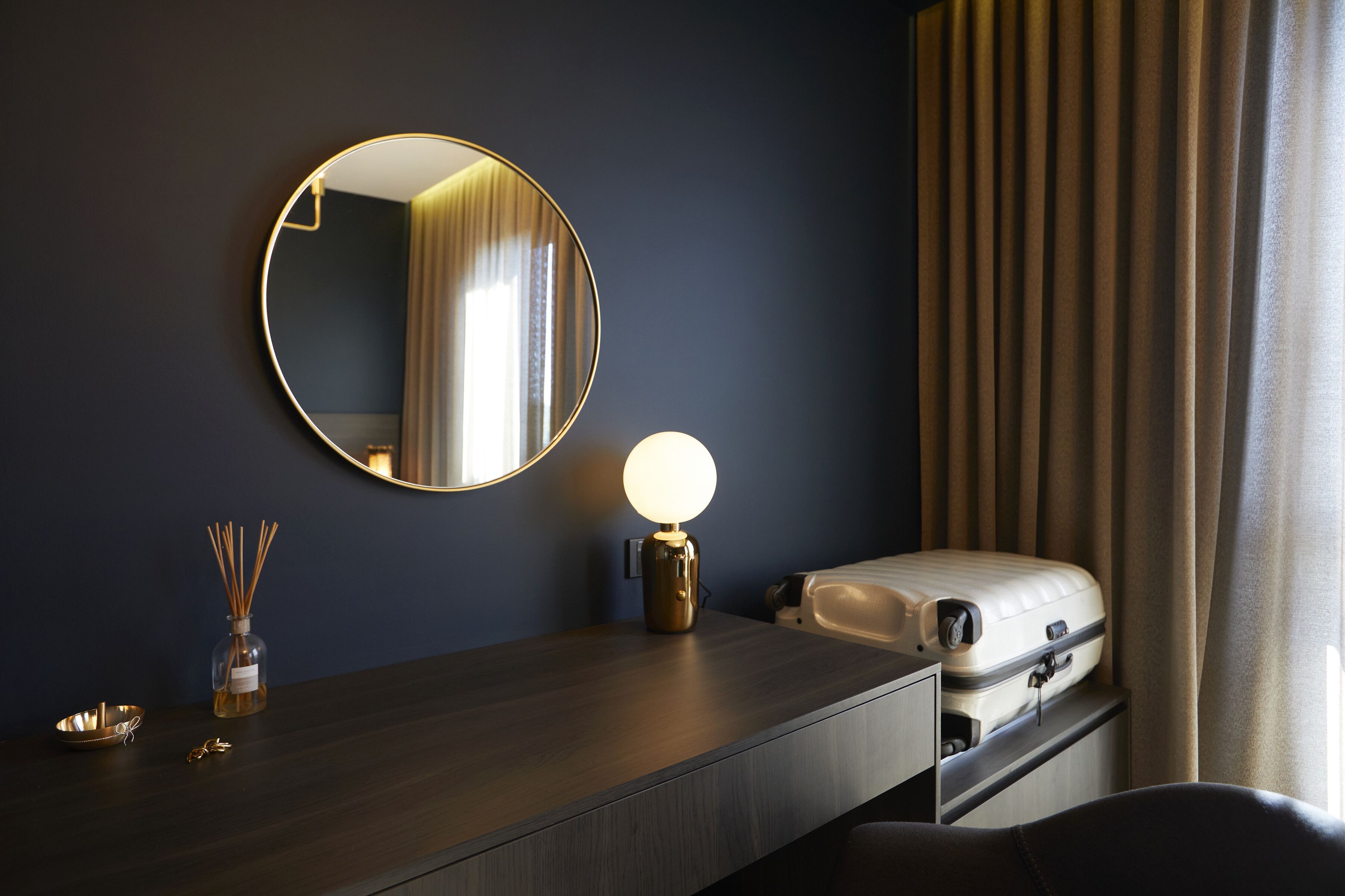 A dark wooden desk with a round mirror, a wall-mounted lamp, a small bottle of reed diffuser, and a suitcase.