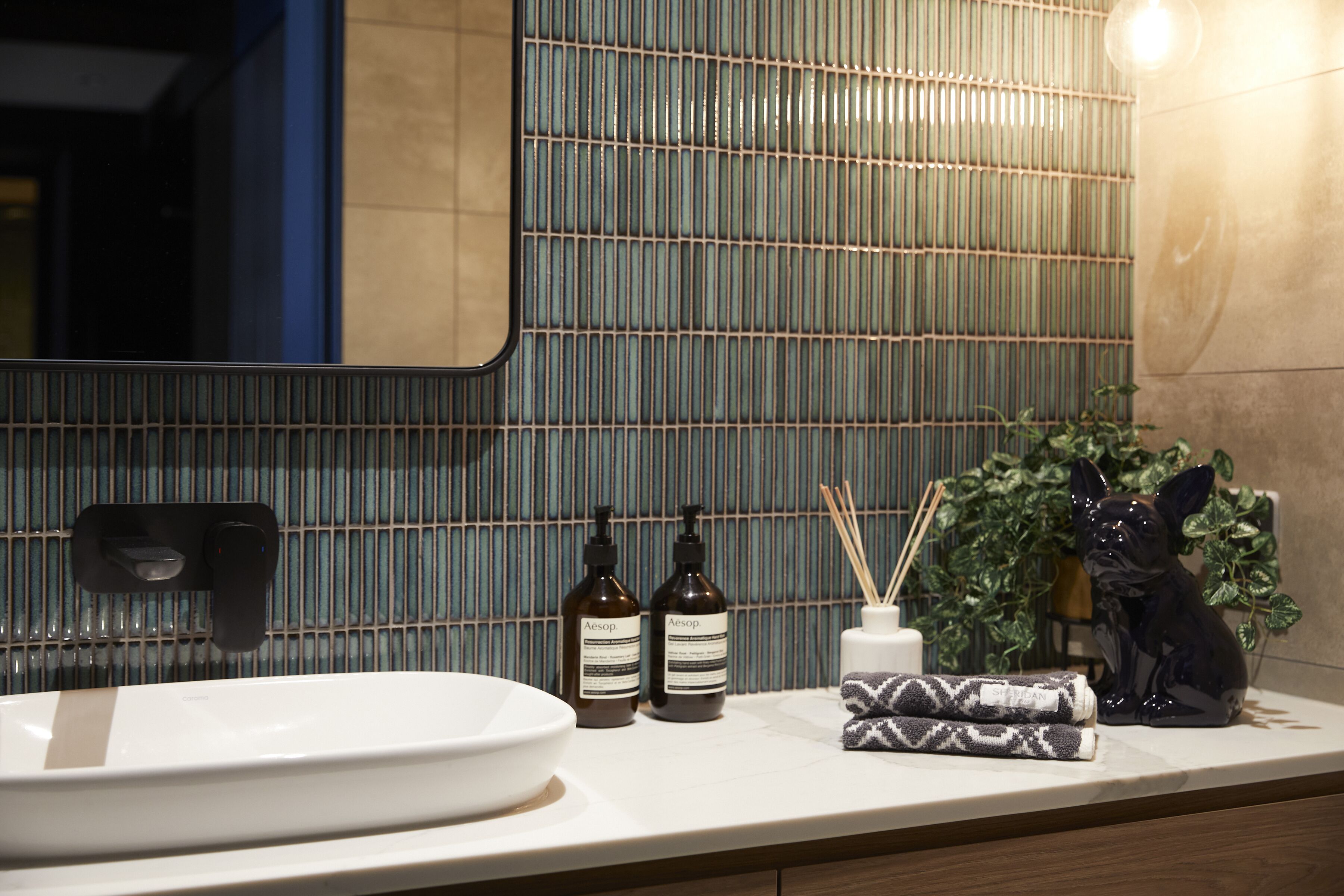 A bathroom with a white sink, two bottles of Aesop shampoo, a black soap dispenser, a decorative fox figurine, and a potted plant.