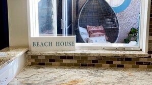 Une cuisine avec des meubles blancs, un plan de travail en marbre et une fenêtre donnant sur une maison de plage.