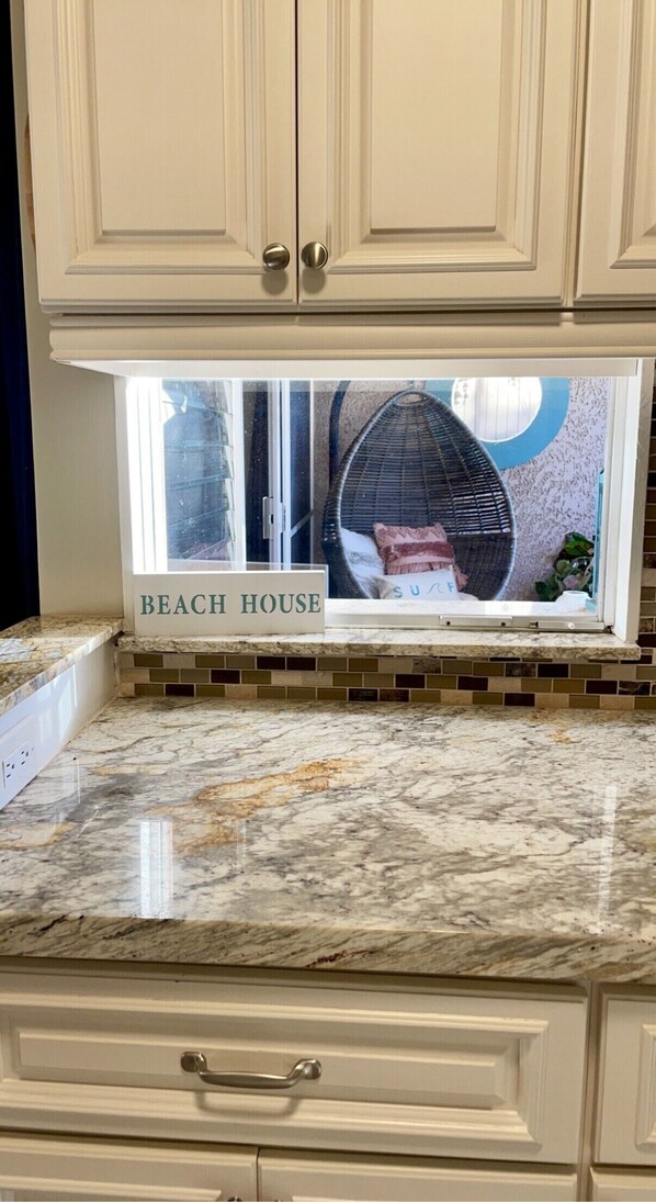 Cucina con mobili bianchi, un piano di lavoro in marmo e una finestra vista su una casa sulla spiaggia.