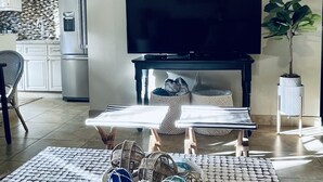 Un salon avec un téléviseur à écran plat, une table basse agrémentée d’objets décoratifs, une plante en pot et, au fond, une cuisine.