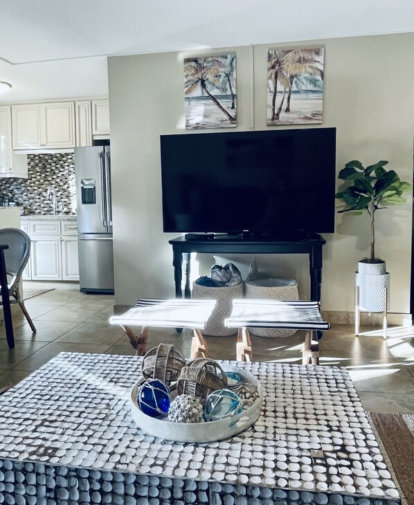 Een woonkamer met een flatscreen televisie, een bijzettafeltje met decoratieve objecten, een potplant en een keuken op de achtergrond.