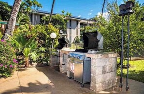 An outdoor barbecue area with a grill and a trash bin, surrounded by greenery and a house in the background. - Kihei Surf Cove Ocean +Jeep (Kihei)