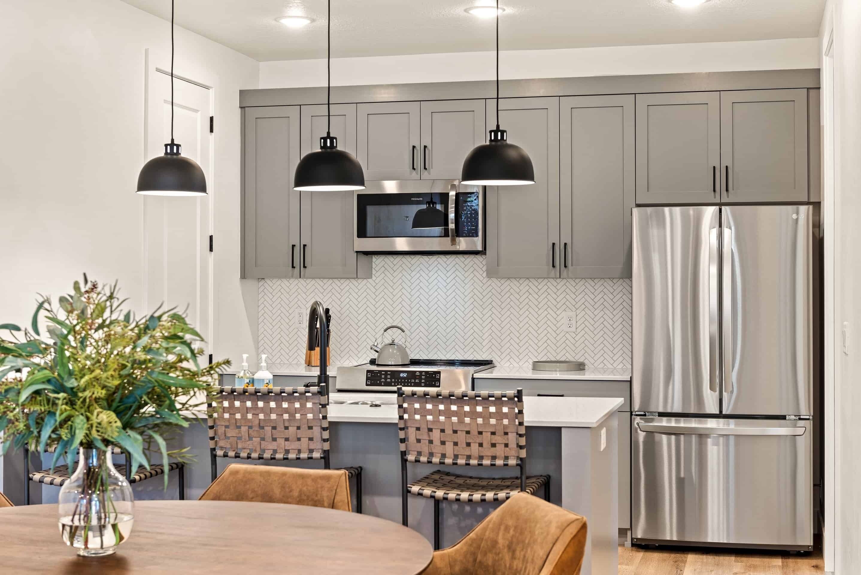 A modern kitchen with grey cabinets, a stainless steel refrigerator, a microwave, and a stove. There are two pendant lights, a vase with greenery on the dining table, and woven chairs.