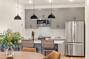 A modern kitchen with grey cabinets, a stainless steel refrigerator, a microwave, and a stove. There are two pendant lights, a vase with greenery on the dining table, and woven chairs.