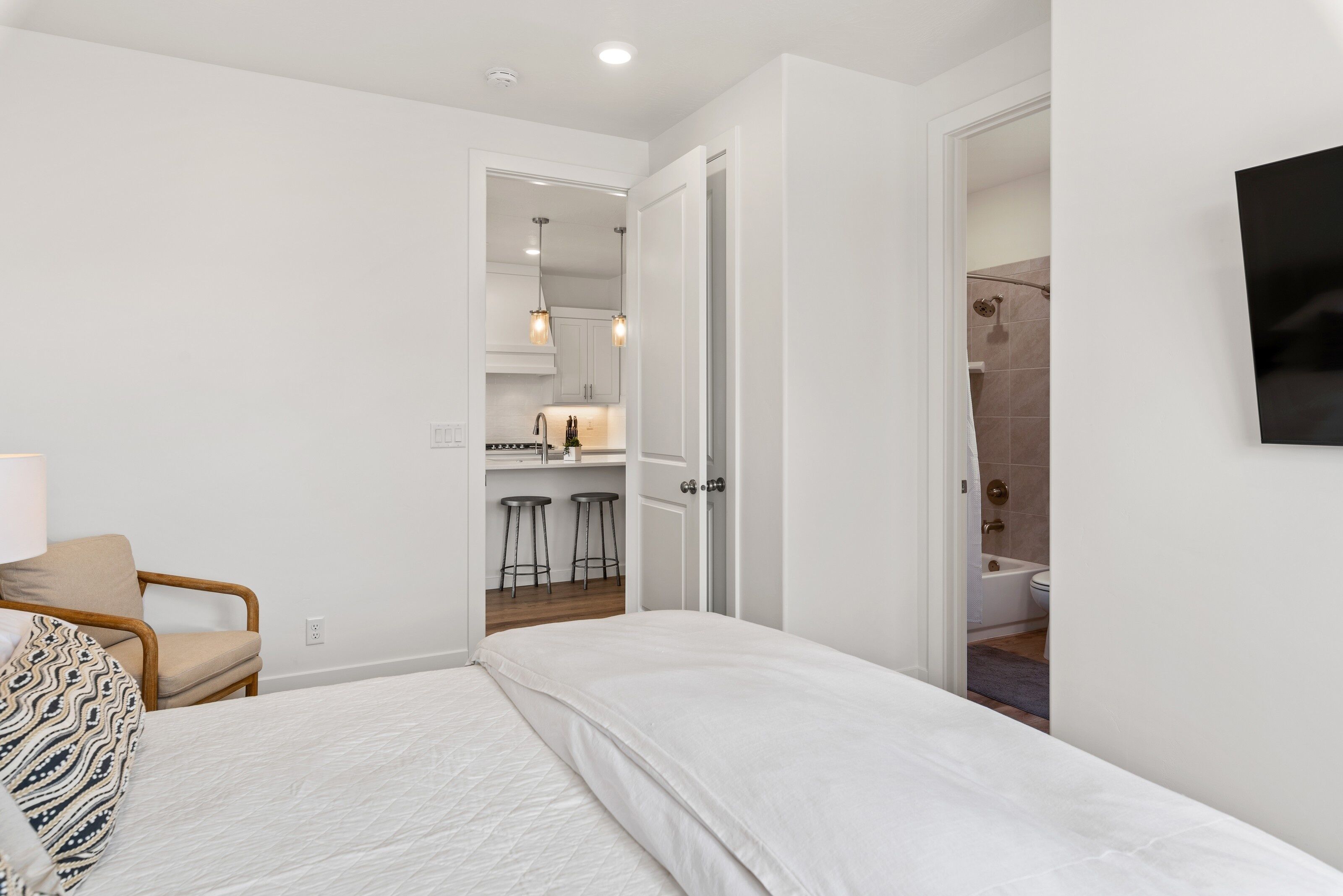 A bedroom with a bed, a chair, and a view into the kitchen and bathroom.