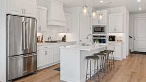 A modern kitchen with white cabinetry, stainless steel appliances, and a central island with bar stools. - The Berber at Desert Color by RedAwning (St. George)