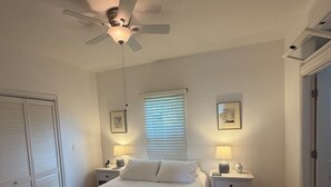 Une chambre à coucher avec un lit, deux tables de chevet avec des lampes, un ventilateur de plafond, une fenêtre avec des stores et une porte dotée d’une petite fenêtre.