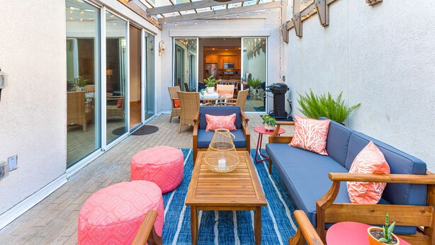 Un patio exterior con una mesa de madera, asientos en tonos azul y rosa, y una cocina visible a través de puertas de vidrio.