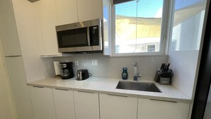 A modern kitchen with white cabinets, a stainless steel microwave, a coffee maker, a toaster, a sink, and a window with a view of a building outside. - Modern studio fully equipped (Doral)