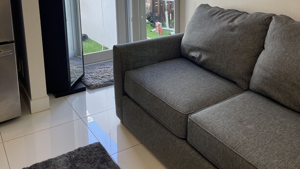 A living room with a grey sofa, a black cabinet, and a sliding glass door leading to a balcony with a view of a building and a garden. - Modern studio fully equipped (Doral)