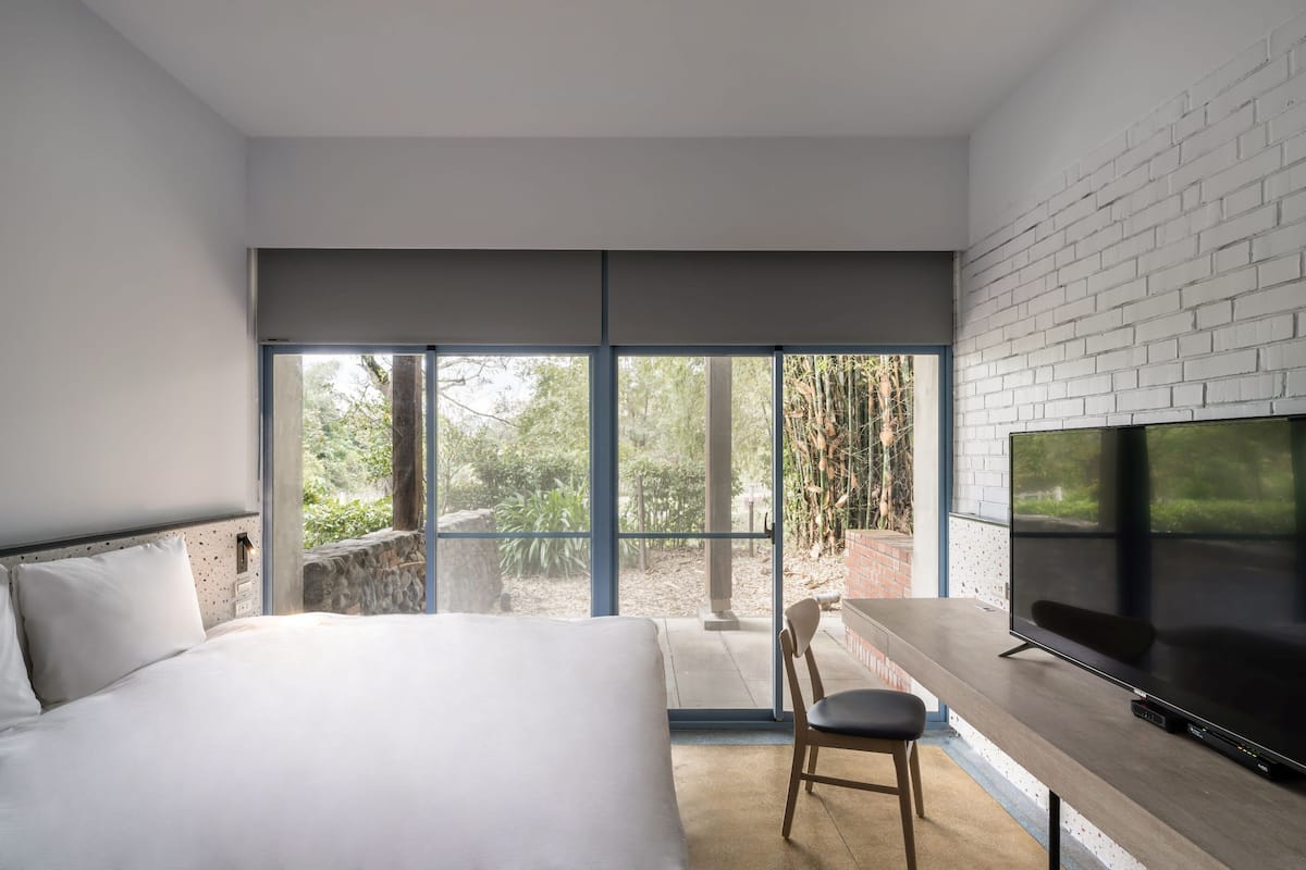 A modern hotel room with a large bed, a TV, and a balcony with a view of greenery.