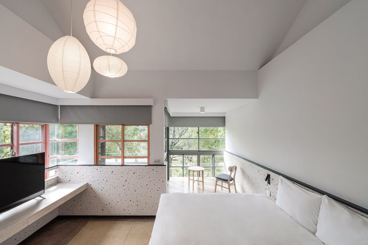 A modern bedroom with a large bed, a TV on a built-in shelf, a small table, and a chair. There are large windows with a view of trees.