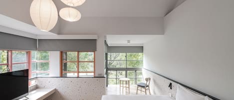 Un dormitorio moderno con una cama grande, un televisor en un estante empotrado, una mesita y una silla. Amplias ventanas con vistas a árboles.