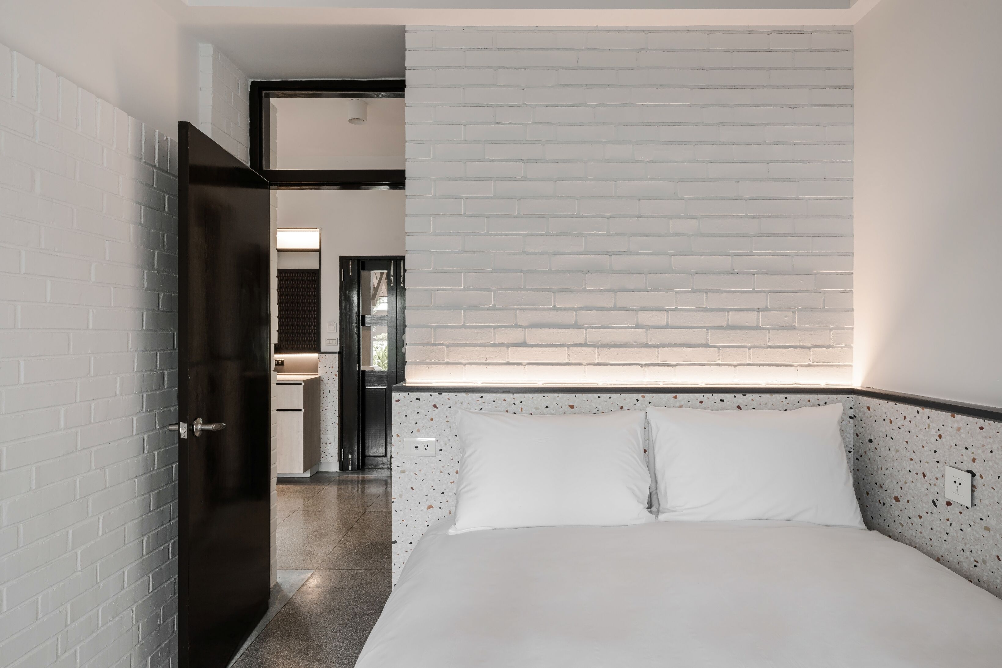 A modern hotel room with a white brick wall, a bed with white bedding, and a small kitchenette in the background.
