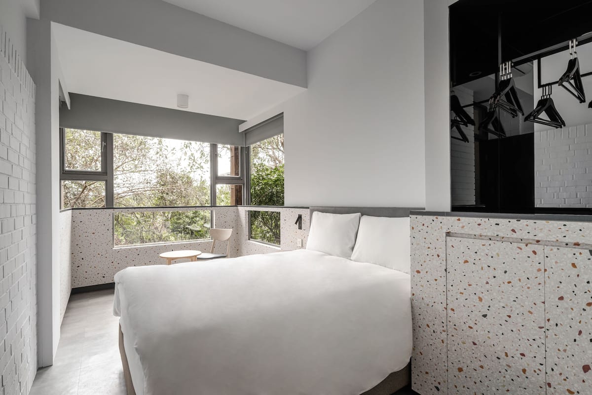 A modern bedroom with a large bed, a small round table, and a balcony with a view of trees.