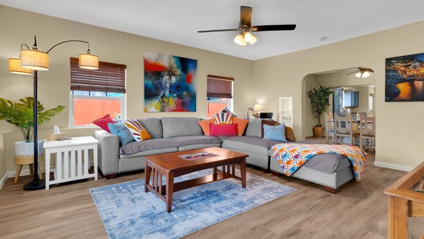 A spacious living room with a gray sofa, a wooden coffee table, and a blue rug. There are two ceiling fans, a floor lamp, and a large abstract painting on the wall. - Midtown Territorial Home Beautifully Updated + Convenient  2 miles U of A (Tucson)